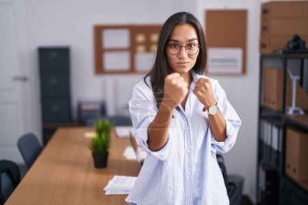 Photo for Young hispanic woman at the office ready to fight with fist defense gesture, angry and upset face, afraid of problem - Royalty Free Image