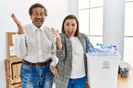 Foto de Pareja interracial de mediana edad sosteniendo la papelera de reciclaje con botellas de plástico en la oficina celebrando la victoria con sonrisa feliz y expresión ganadora con las manos levantadas - Imagen libre de derechos