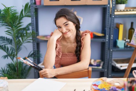 Téléchargez les photos : Jeune artiste hispanique souriante confiante tenant des pinceaux au studio d'art - en image libre de droit