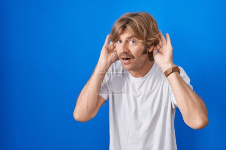 Photo for Middle age man standing over blue background trying to hear both hands on ear gesture, curious for gossip. hearing problem, deaf - Royalty Free Image