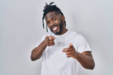 Foto de Hombre africano con rastas con camiseta casual sobre fondo blanco señalando los dedos a la cámara con cara alegre y divertida. buena energía y vibraciones. - Imagen libre de derechos
