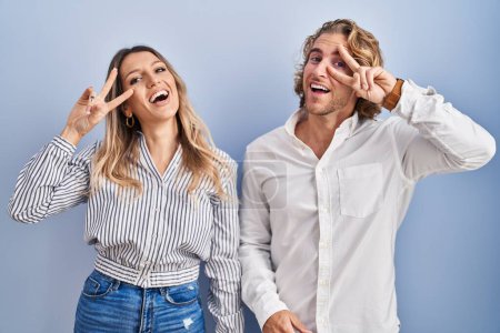 Téléchargez les photos : Jeune couple debout sur fond bleu faisant symbole de paix avec les doigts sur le visage, souriant gai montrant la victoire - en image libre de droit