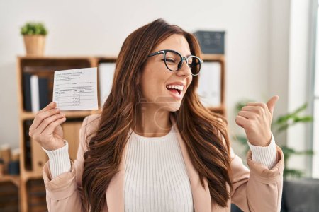 Téléchargez les photos : Jeune femme brune tenant une carte de disque covid pointant le pouce vers le côté souriant heureux avec la bouche ouverte - en image libre de droit
