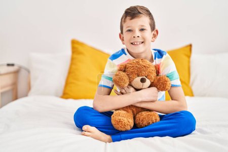 Photo for Blond child hugging teddy bear sitting on bed at bedroom - Royalty Free Image