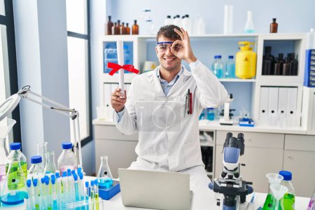 Téléchargez les photos : Young hispanic man working at scientist laboratory holding diploma smiling happy doing ok sign with hand on eye looking through fingers - en image libre de droit