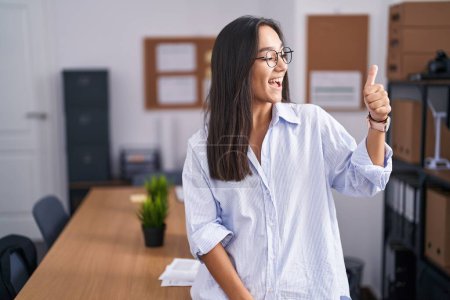 Photo for Young hispanic woman at the office looking proud, smiling doing thumbs up gesture to the side - Royalty Free Image