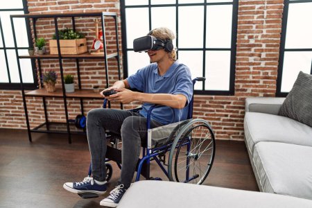 Foto de Joven hombre rubio jugando videojuego con gafas de realidad virtual y joystick sentado en silla de ruedas en casa - Imagen libre de derechos