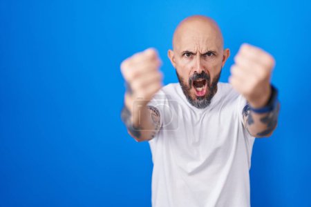 Téléchargez les photos : Homme hispanique avec des tatouages debout sur fond bleu en colère et fou levant les poings frustrés et furieux tout en criant de colère. rage et concept agressif. - en image libre de droit