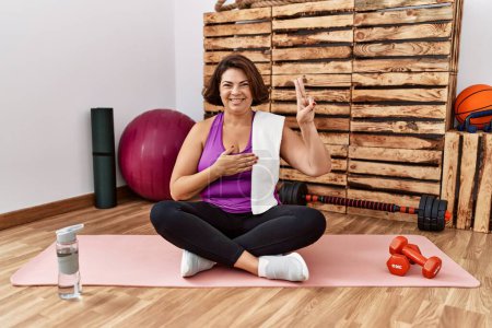Foto de Middle age hispanic woman sitting on training mat at the gym smiling swearing with hand on chest and fingers up, making a loyalty promise oath - Imagen libre de derechos