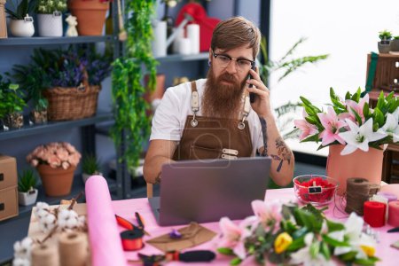 Photo for Young redhead man florist talking on smartphone using laptop at flower shop - Royalty Free Image