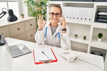 Photo for Young doctor woman speaking on the phone doing ok sign with fingers, smiling friendly gesturing excellent symbol - Royalty Free Image