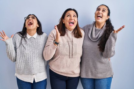 Photo for Mother and two daughters standing over blue background crazy and mad shouting and yelling with aggressive expression and arms raised. frustration concept. - Royalty Free Image