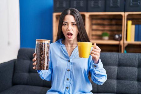 Téléchargez les photos : Jeune femme brune tenant des grains de café et une tasse de café dans le visage choqué, l'air sceptique et sarcastique, surpris avec la bouche ouverte - en image libre de droit