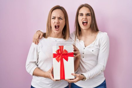 Foto de Middle age mother and young daughter holding mothers day gift angry and mad screaming frustrated and furious, shouting with anger. rage and aggressive concept. - Imagen libre de derechos