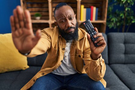 Téléchargez les photos : African american man holding television remote control with open hand doing stop sign with serious and confident expression, defense gesture - en image libre de droit