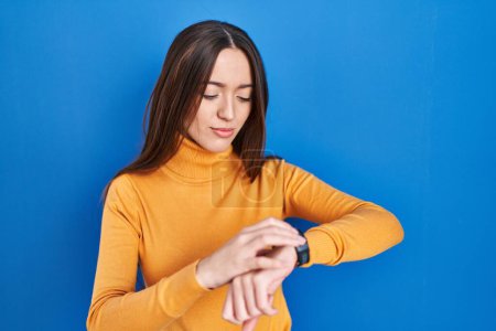 Téléchargez les photos : Jeune femme brune debout sur fond bleu vérifier l'heure sur montre-bracelet, détendu et confiant - en image libre de droit