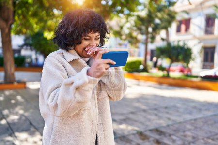 Foto de Joven hermosa mujer hispana sonriendo confiada viendo video en smartphone en el parque - Imagen libre de derechos
