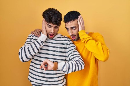 Téléchargez les photos : Jeune couple gay hispanique debout sur fond jaune regardant le temps de la montre inquiet, peur d'arriver en retard - en image libre de droit