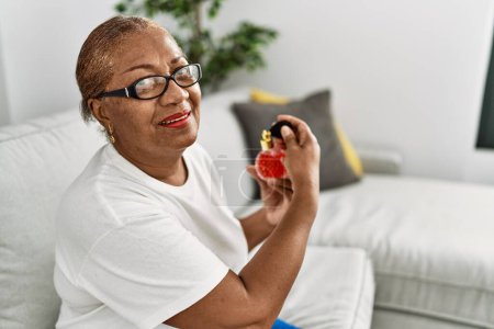 Téléchargez les photos : Femme afro-américaine senior utilisant un parfum assis sur le canapé à la maison - en image libre de droit