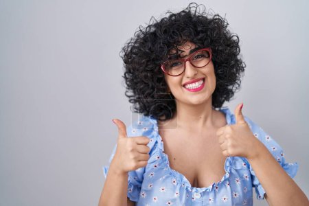 Téléchargez les photos : Young brunette woman with curly hair wearing glasses over isolated background success sign doing positive gesture with hand, thumbs up smiling and happy. cheerful expression and winner gesture. - en image libre de droit