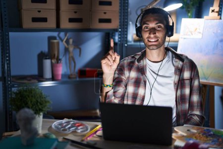 Foto de Joven hombre hispano sentado en el estudio de arte con portátil tarde en la noche mostrando y señalando con el dedo número uno mientras sonríe confiado y feliz. - Imagen libre de derechos