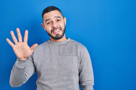 Foto de Hispanic man standing over blue background showing and pointing up with fingers number five while smiling confident and happy. - Imagen libre de derechos