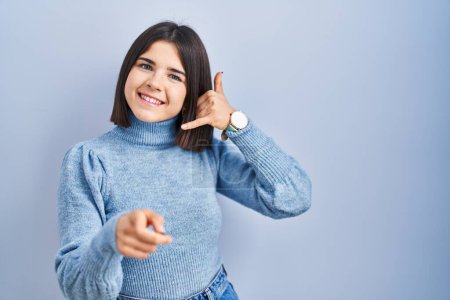 Photo for Young hispanic woman standing over blue background smiling doing talking on the telephone gesture and pointing to you. call me. - Royalty Free Image