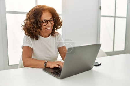 Foto de Mujer hispana de mediana edad sonriendo confiada usando laptop en casa - Imagen libre de derechos