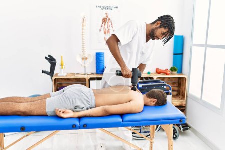 Foto de Dos hombres fisioterapeuta y paciente masajeando la espalda usando pistola de percusión en la clínica - Imagen libre de derechos
