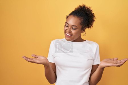 Téléchargez les photos : Young hispanic woman with curly hair standing over yellow background smiling showing both hands open palms, presenting and advertising comparison and balance - en image libre de droit