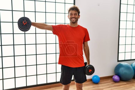 Téléchargez les photos : Jeune homme hispanique souriant entraînement confiant à l'aide d'haltères au centre sportif - en image libre de droit