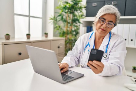Téléchargez les photos : Middle age woman wearing doctor uniform using smartphone at clinic - en image libre de droit