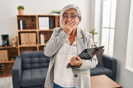 Photo for Middle age woman with grey hair at consultation office laughing and embarrassed giggle covering mouth with hands, gossip and scandal concept - Royalty Free Image