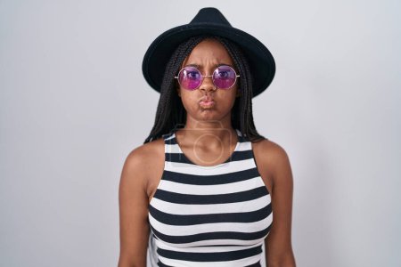 Photo for Young african american with braids wearing hat and sunglasses puffing cheeks with funny face. mouth inflated with air, crazy expression. - Royalty Free Image