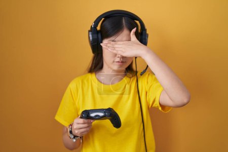 Photo for Chinese young woman playing video game holding controller covering eyes with hand, looking serious and sad. sightless, hiding and rejection concept - Royalty Free Image