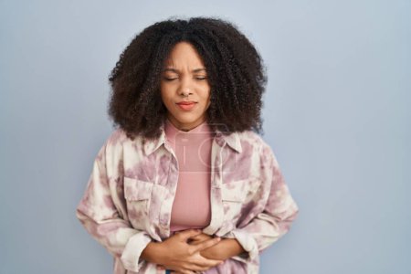 Téléchargez les photos : Jeune femme afro-américaine debout sur fond bleu avec la main sur le ventre parce que les nausées, la maladie douloureuse se sentent mal. concept de douleur. - en image libre de droit