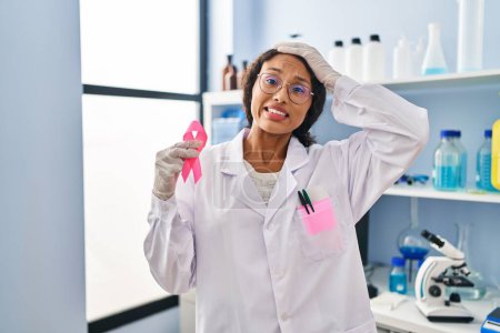 Photo for Young hispanic doctor woman working at scientist laboratory holding pink ribbon stressed and frustrated with hand on head, surprised and angry face - Royalty Free Image
