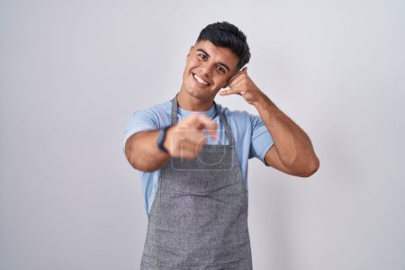 Photo for Hispanic young man wearing apron over white background smiling doing talking on the telephone gesture and pointing to you. call me. - Royalty Free Image