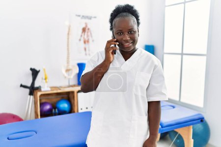 Foto de Young african american woman wearing physiotherapist uniform talking on the smartphone at clinic - Imagen libre de derechos