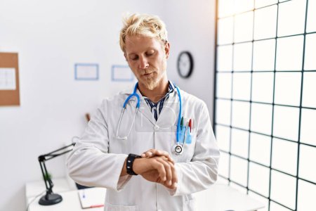 Foto de Joven hombre rubio vistiendo uniforme médico y estetoscopio en la clínica comprobar la hora en el reloj de pulsera, relajado y seguro de sí mismo - Imagen libre de derechos