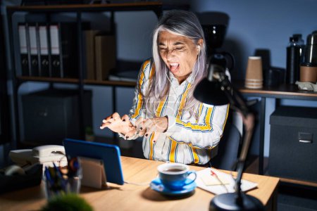 Foto de Mujer de mediana edad con el pelo gris trabajando en la oficina por la noche expresión disgustada, disgustada y temerosa haciendo asqueroso rostro porque la reacción de aversión. con las manos levantadas - Imagen libre de derechos