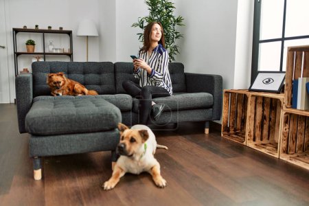 Photo for Young hispanic woman using smartphone sitting on sofa with dogs at home - Royalty Free Image