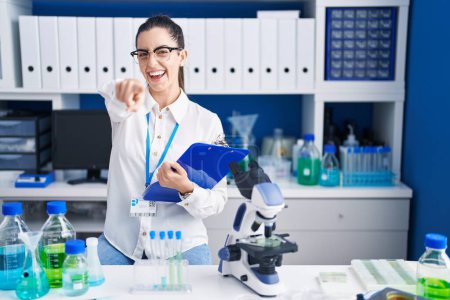 Photo for Young brunette woman working at scientist laboratory pointing to you and the camera with fingers, smiling positive and cheerful - Royalty Free Image