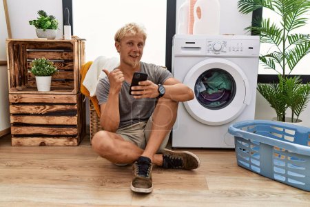 Foto de Young blond man doing laundry using smartphone pointing to the back behind with hand and thumbs up, smiling confident - Imagen libre de derechos