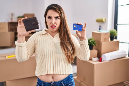 Téléchargez les photos : Young hispanic woman moving to a new home holding wallet and credit card clueless and confused expression. doubt concept. - en image libre de droit