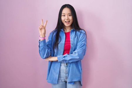 Photo for Young asian woman standing over pink background smiling with happy face winking at the camera doing victory sign with fingers. number two. - Royalty Free Image