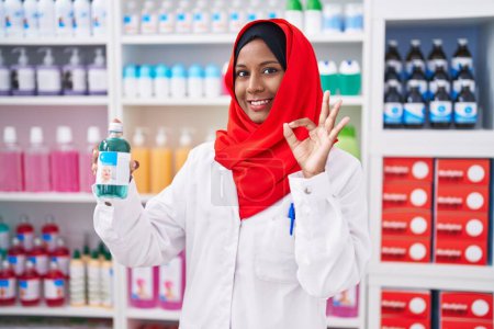Téléchargez les photos : Young arab woman working at pharmacy drugstore holding syrup doing ok sign with fingers, smiling friendly gesturing excellent symbol - en image libre de droit