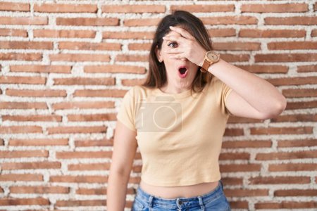 Photo for Young brunette woman standing over bricks wall peeking in shock covering face and eyes with hand, looking through fingers with embarrassed expression. - Royalty Free Image
