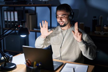 Foto de Joven hombre guapo trabajando con computadora portátil en la noche mostrando y apuntando hacia arriba con los dedos número ocho mientras sonríe confiado y feliz. - Imagen libre de derechos