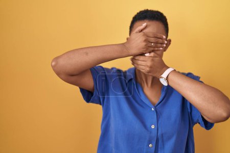 Foto de Mujer afroamericana de pie sobre fondo amarillo cubriendo los ojos y la boca con las manos, sorprendida y conmocionada. ocultar emociones - Imagen libre de derechos